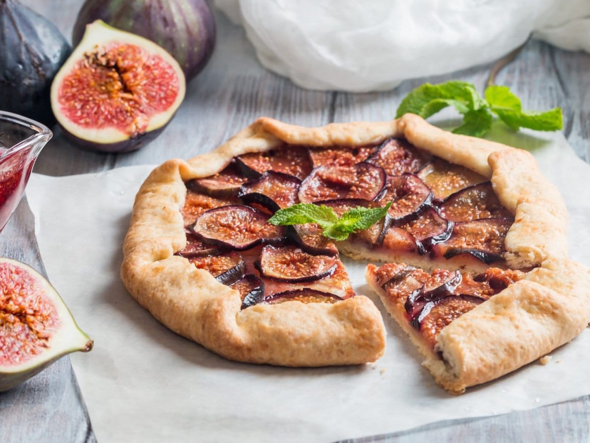 Eine angeschnittene einfache Feigen-Galette auf einem Stück Backpapier, daneben ein Messer, frische, zum Teil aufgeschnittene Feigen und ein Krug mit Feigenmarmelade.