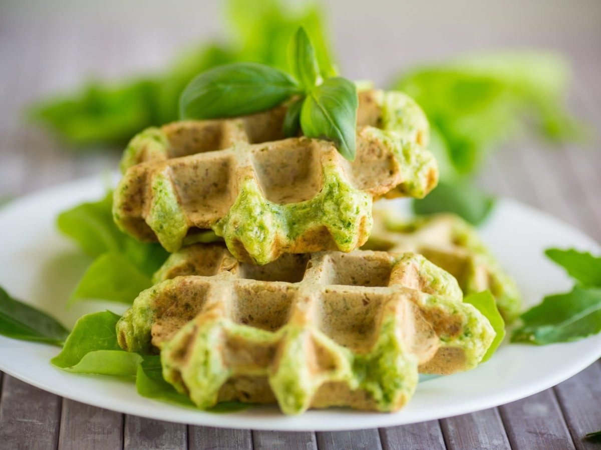 Frontalsicht. Zwei Falafel-Waffeln auf einem weißen Teller. Drum herum liegen frische Kräuter.