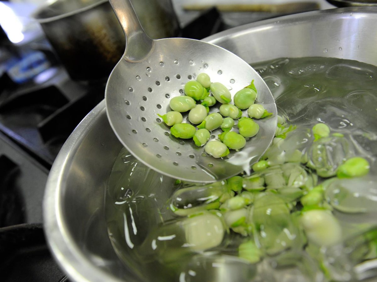 Draufsicht, Nahaufnahme: Eine gelöcherte Küchenkelle auf der Bohnen sind werden in eine Schüssel mit Eiswasser gegeben.