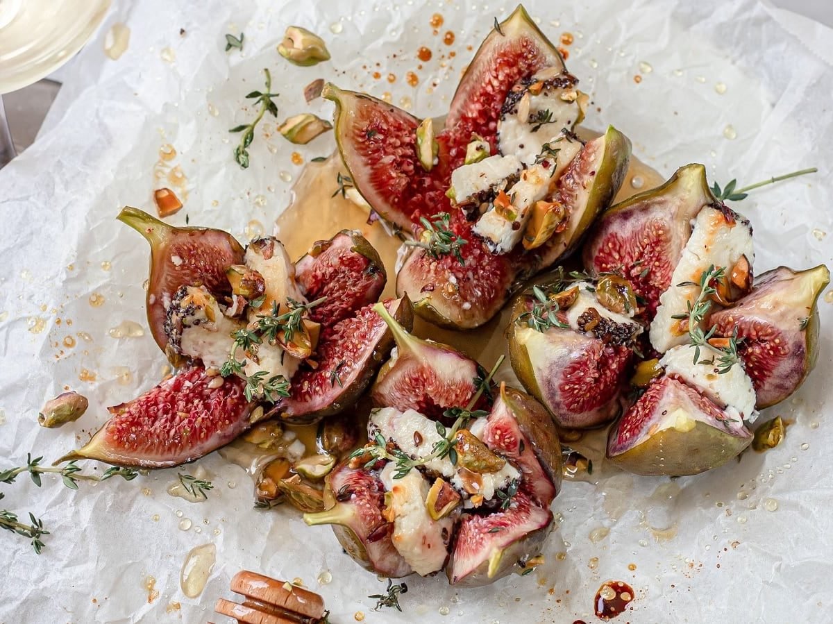 Gebackene Feigen mit Ziegenkäse auf weißem Backpapier. Links unten eine kleine Schüssel mit Pistazien und links oben ein Glas Weißwein.