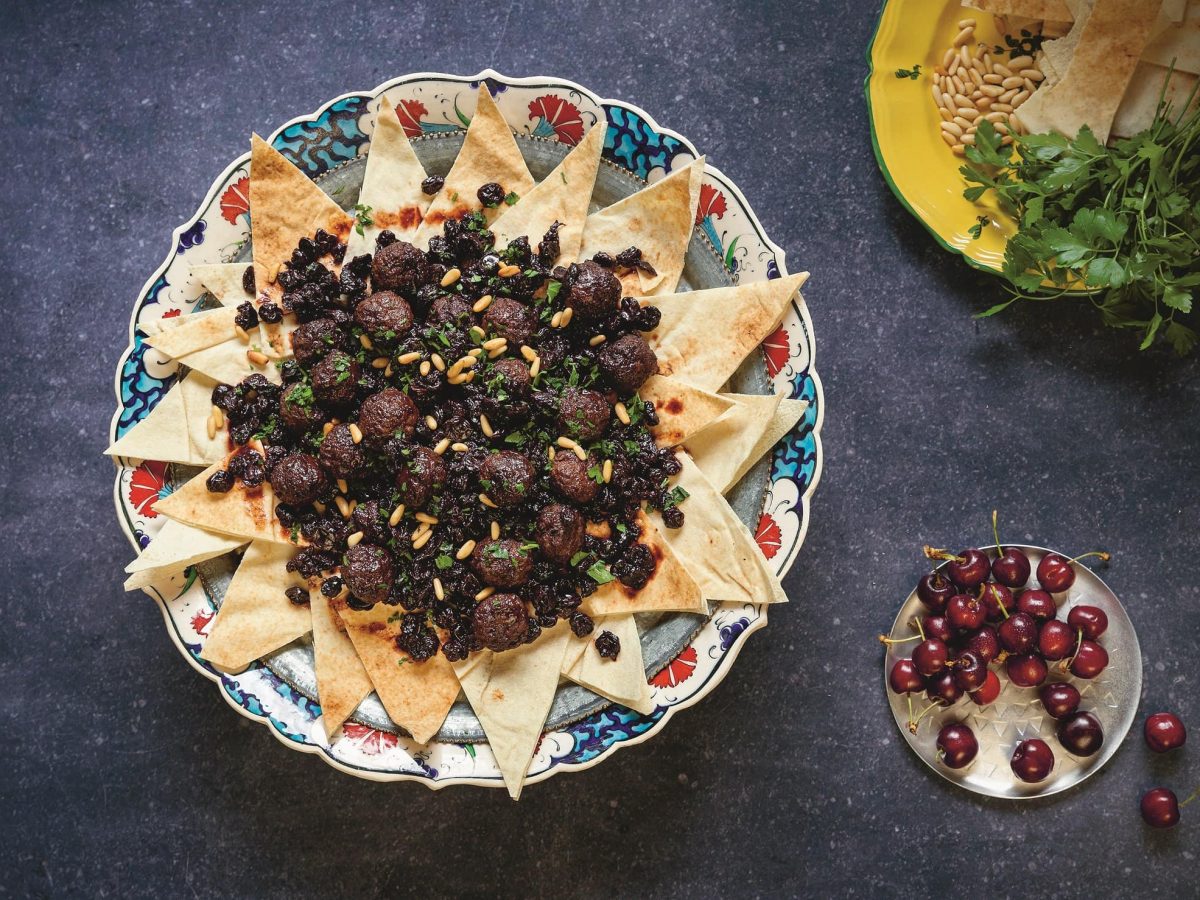 Fleischbällchen mit Sauerkirschen auf in Dreiecke geschnittenen Pitabroten, angerichtet auf einem Teller, Draufsicht, daneben eine Schale Kirschen und Kräuter. Das Rezept aus dem Buch "Granatapfel & Artischocke"