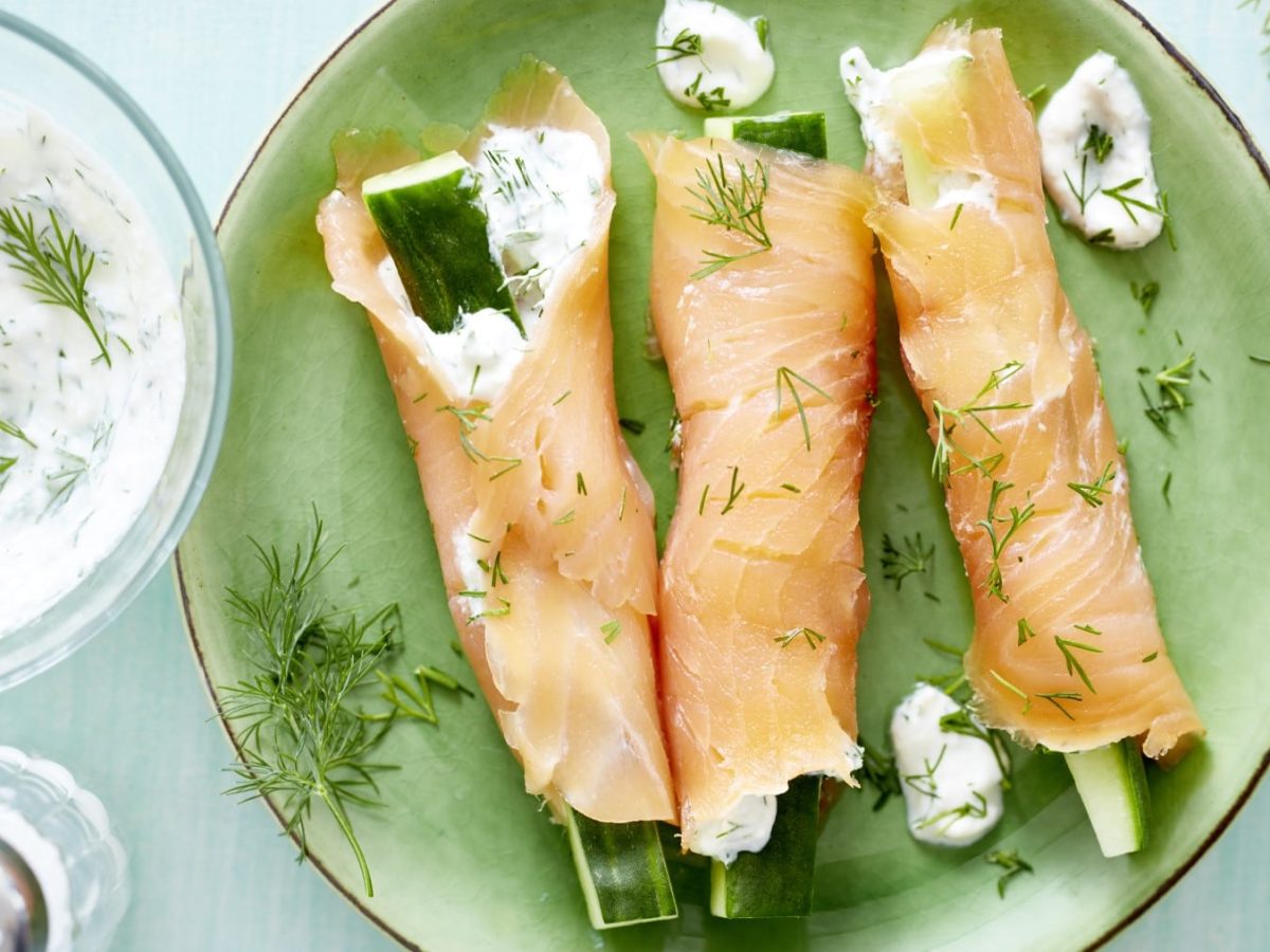 Drei Gefüllte Lachsröllchen auf grünem Teller, daneben weißer Dip mit Dill und Salzstreuer auf hellem Untergrund.