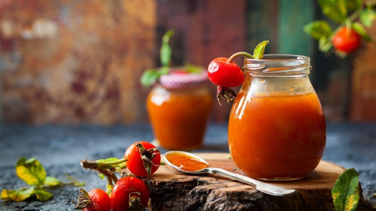 Hagebuttenmarmelade in zwei Gläsern auf blauem Tusch vor Holz. Im Bild Rosenblätter und Hagebutten. Frontalansicht.