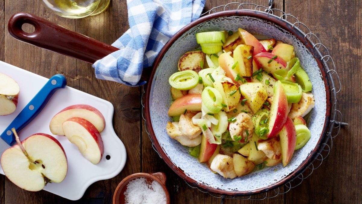 Eine Hähnchen-Kartoffel-Pfanne mit Apfel auf einem hölzernen Hintergrund. Links davon ein Schneidebrett mit Apfel und ein Messer, links unten eine kleine Schüssel mit Salz.