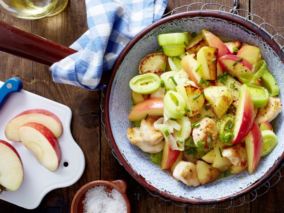 Eine Hähnchen-Kartoffel-Pfanne mit Apfel auf einem hölzernen Hintergrund. Links davon ein Schneidebrett mit Apfel und ein Messer, links unten eine kleine Schüssel mit Salz.