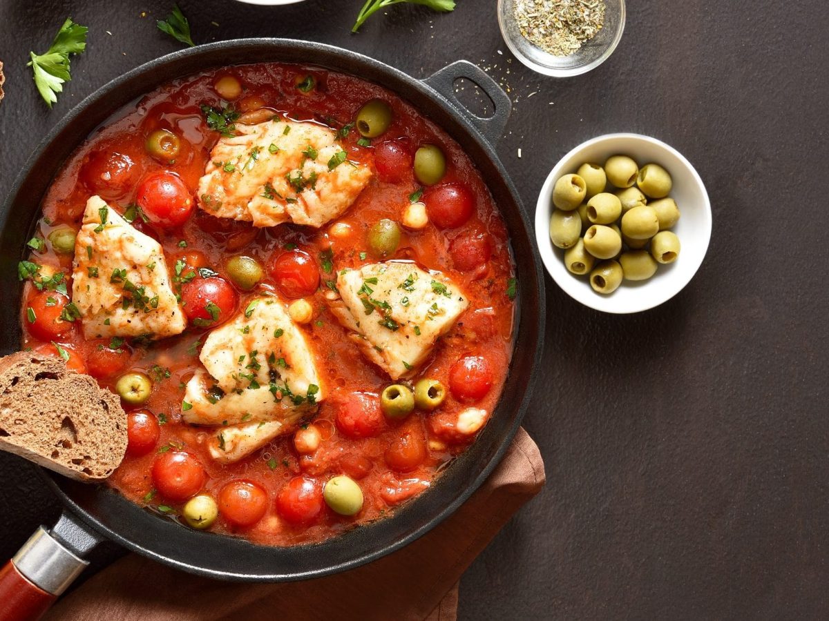 Draufsicht: Eine Kabeljau-Pfanne mit Tomaten, Oliven und Kichererbsen steht auf einem dunklen Untergrund. Drum herum sind Schälchen mit frischen Zutaten und Kräuter.