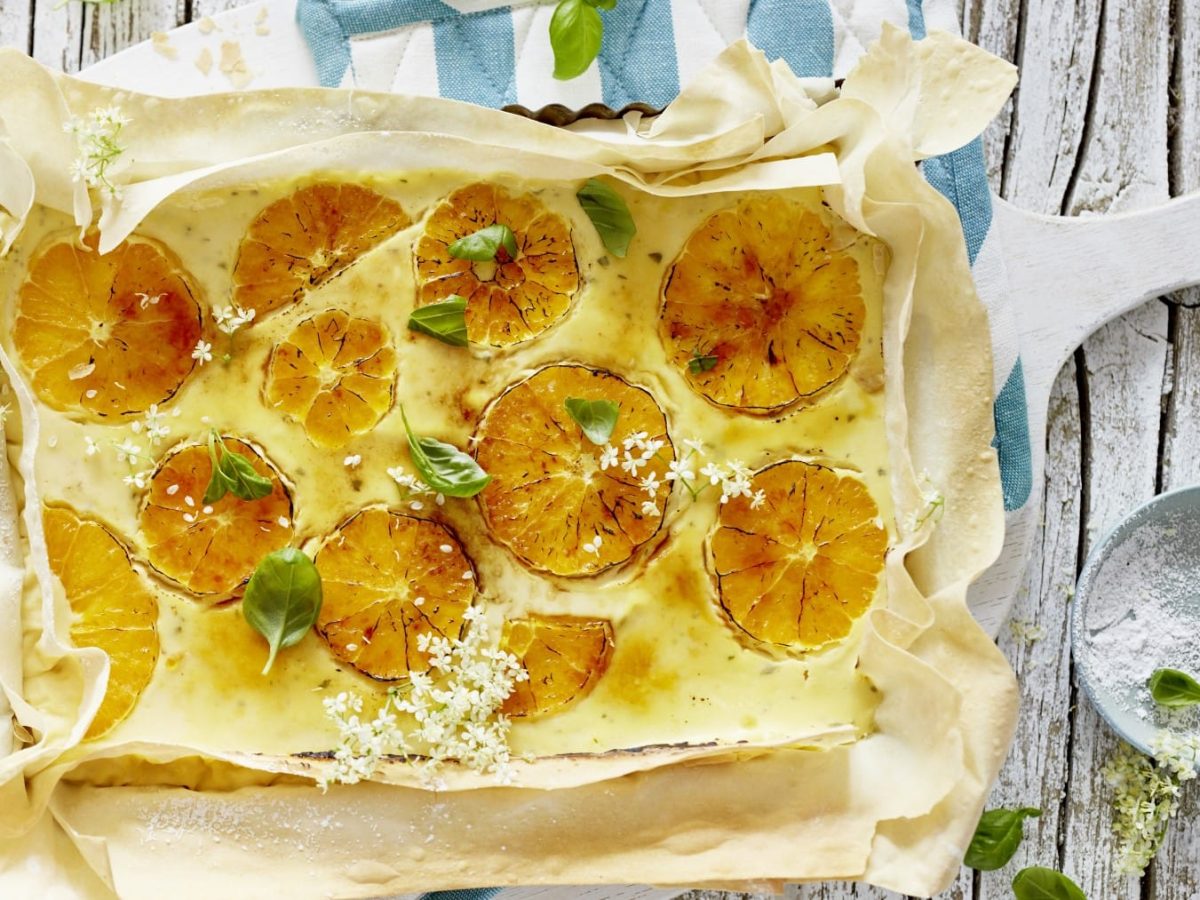 Eine karamellisierte Orangen-Tarte auf blau-weißen Ofentüchern auf einem hölzernen Hintergrund. Rechts davon ein Schälchen mit Blüten und Puderzucker.