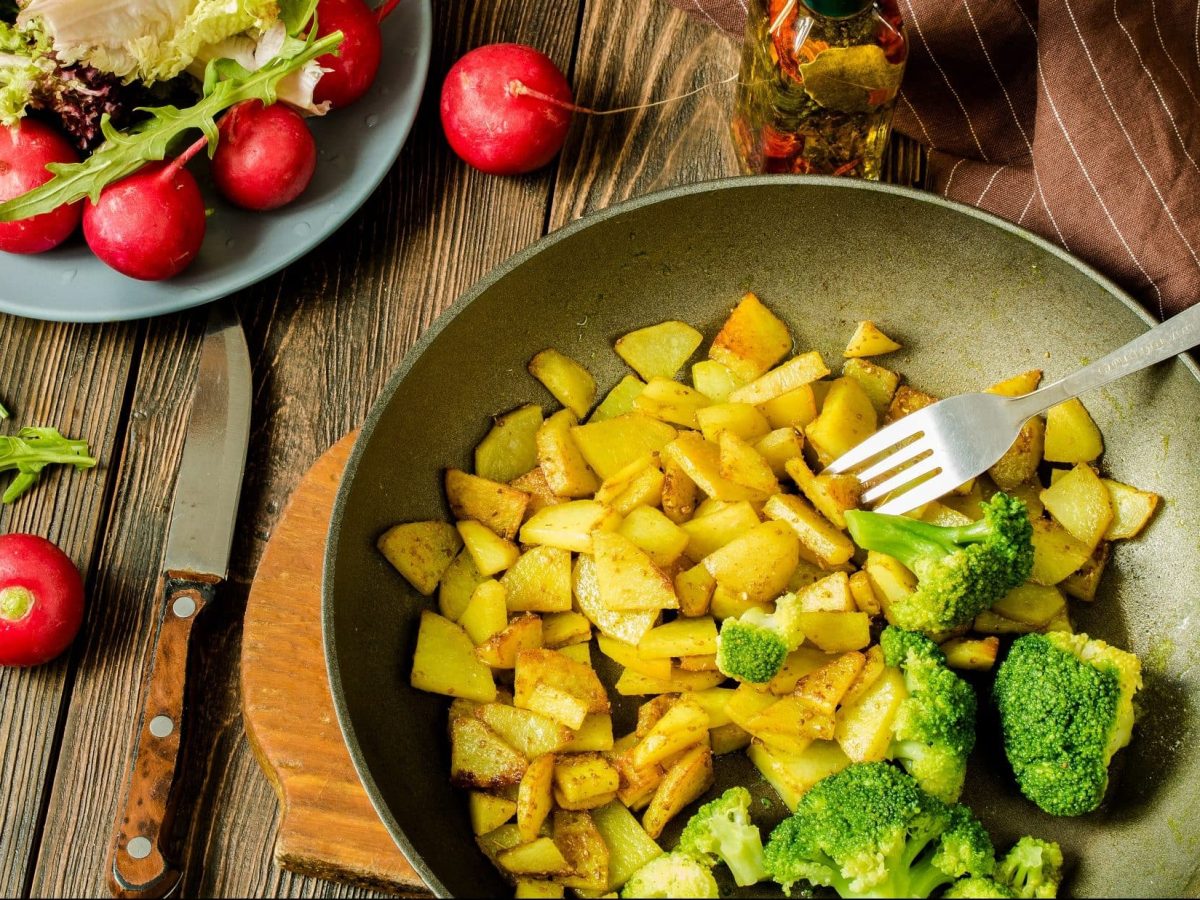 Draufsicht: Untergrund aus Holz, eine Kartoffel-Brokkoli-Pfanne und drum herum liegen Radieschen. Daneben ist eine Schüssel Salat.