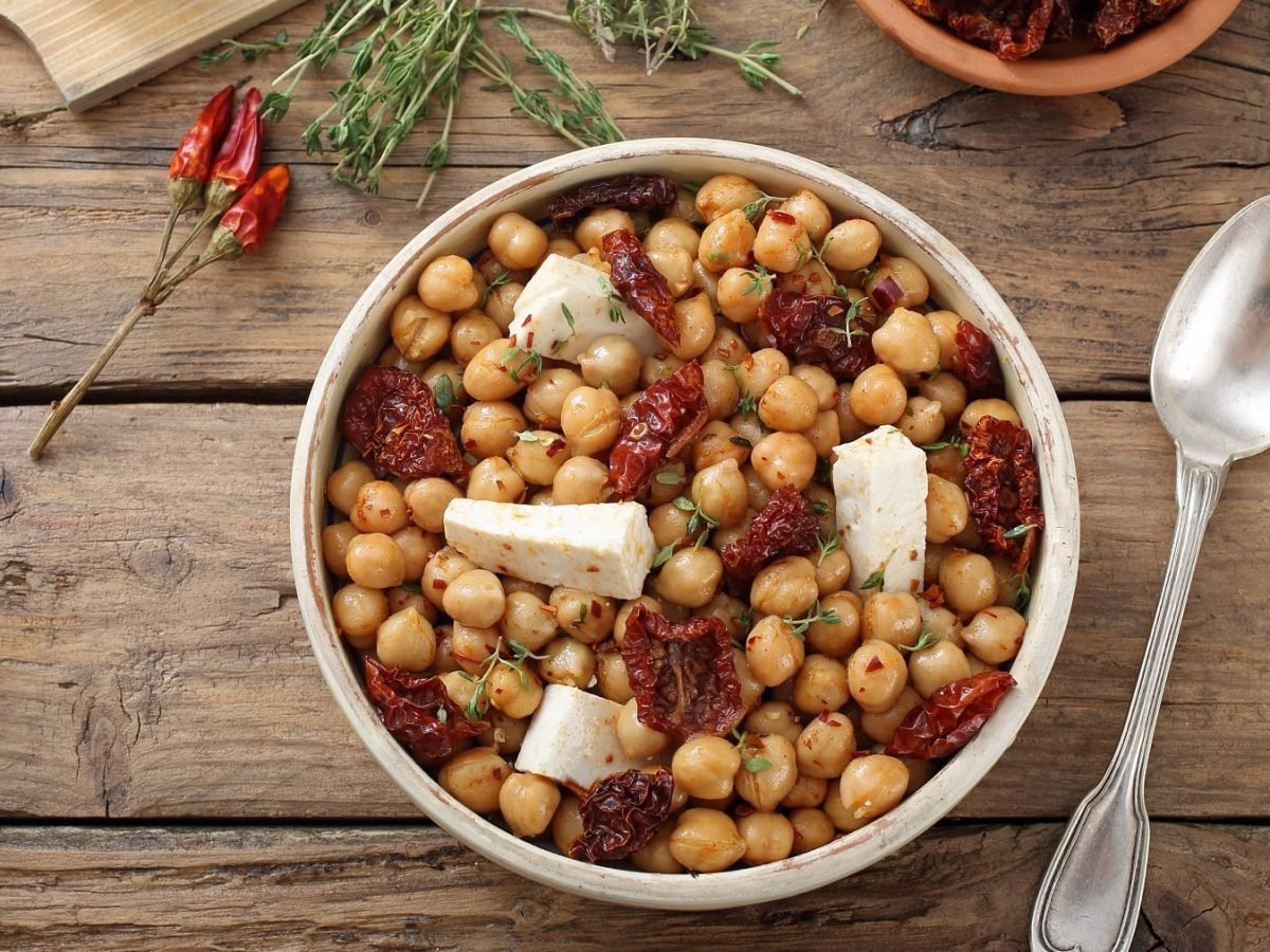 Eine weiße Schale mit dem Salat direkt von oben fotografiert. Darüber Feta, getrocknete Tomaten und Thymian. Alles auf einem rustikalen Holztisch.