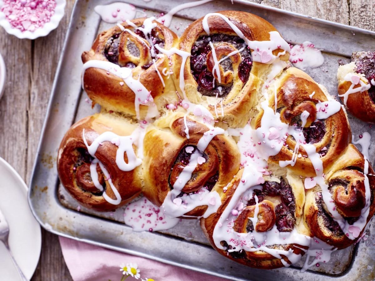 Kirsch-Mohn-Schnecken auf einem silbernen Blech auf einem hölzernen Hintergrund. Links davon ein kleiner Teller mit einer Kirsch-Mohn-Schnecke, eine kleine Schüssel mit Zuckerguss und Zuckerstreusel. Rechts ein rosarotes Geschirrtuch.