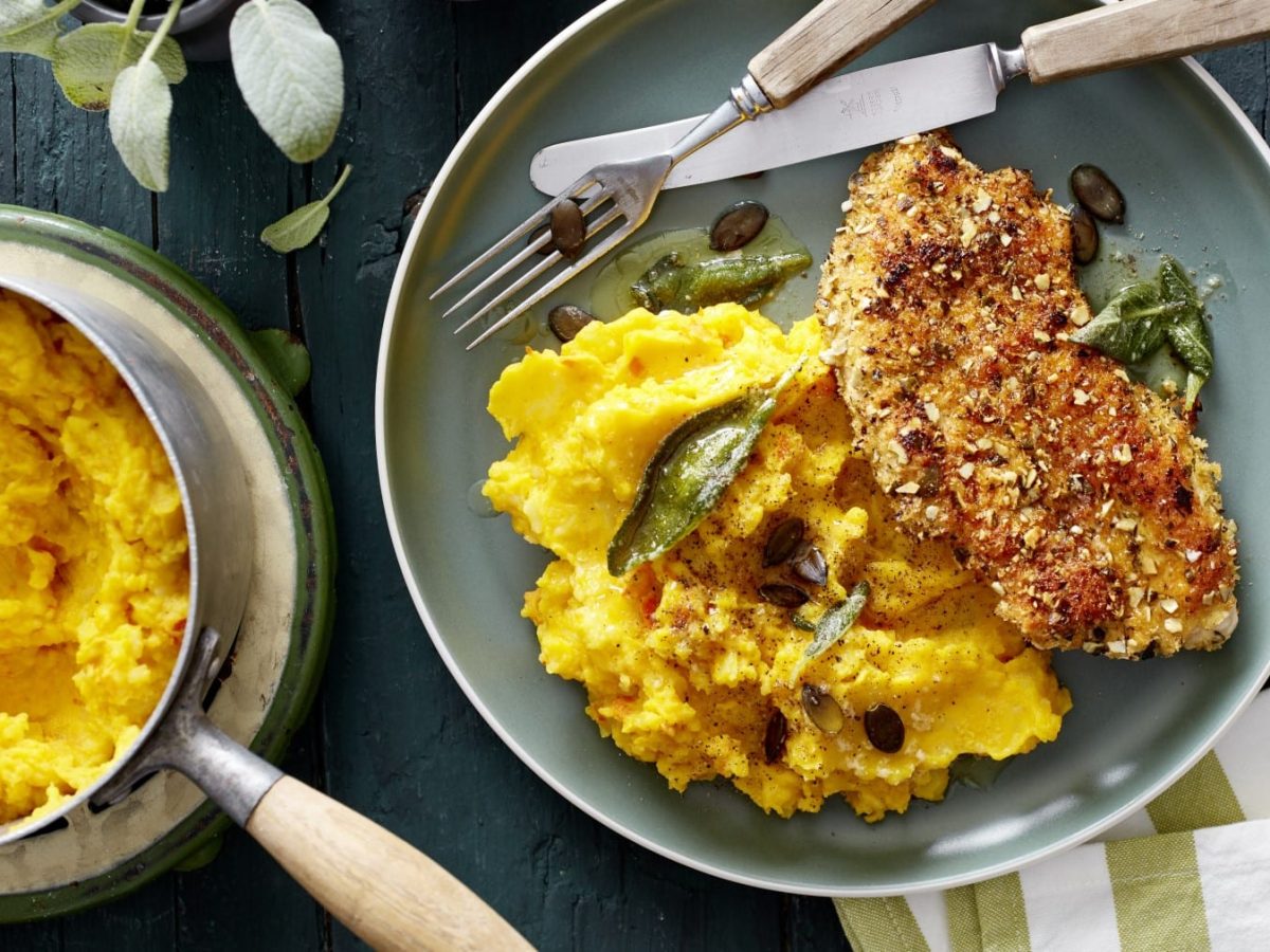 Ein Teller mit Knusper-Schnitzel mit Kürbisstampf und Besteck, daneben Topf mit Kürbisbrei auf dunklem Untergrund.