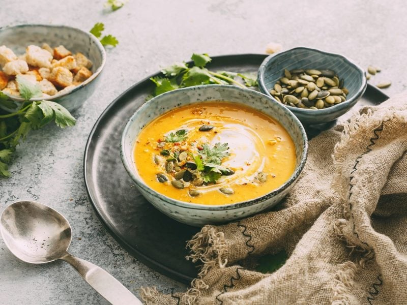 Eine Schüssel Kürbis-Curry-Suppe mit Kürbiskernen, Kräutern und Schmand, daneben ein Löffel, ein Geschirrtuch, eine Schale mit Kürbiskernen und Croûtons.