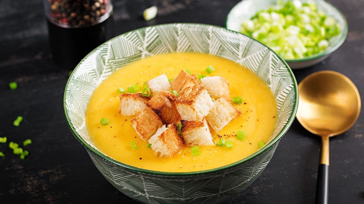 Eine gemusterte Schüssel Kürbis-Kartoffel-Suppe mit Croûtons und Frühlingszwiebeln, daneben ein goldener Löffel und eine Schale mit gehackten Frühlingszwiebeln.