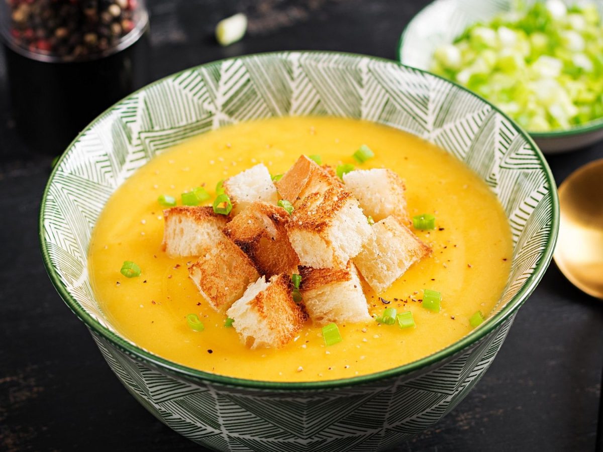 Eine gemusterte Schüssel Kürbis-Kartoffel-Suppe mit Croûtons und Frühlingszwiebeln, daneben ein goldener Löffel und eine Schale mit gehackten Frühlingszwiebeln.