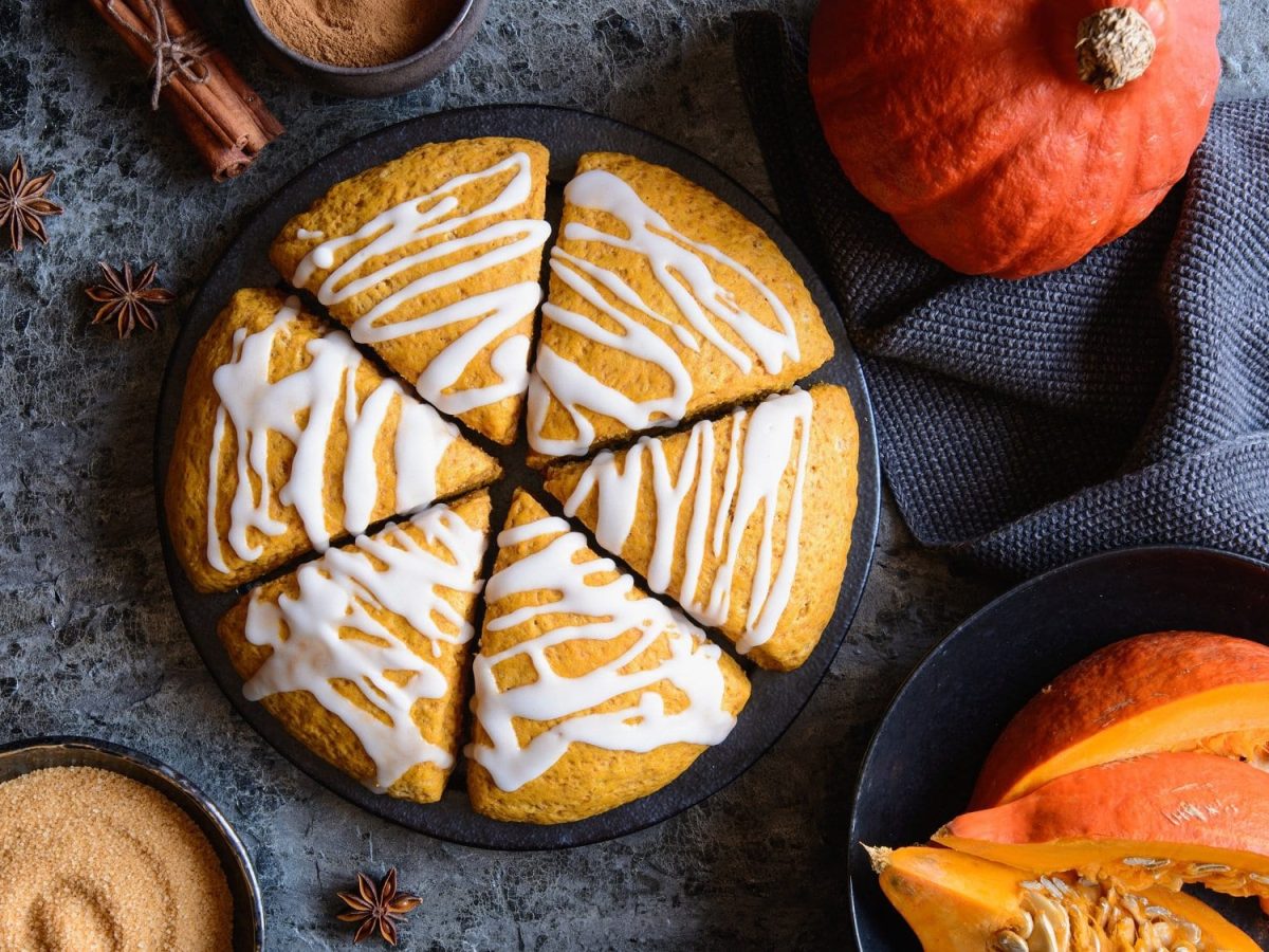 6 Kürbis-Scones wie eine Torte arrangiert auf einem Teller in der Draufsicht, drumherum ein ganzer und ein geschnittener Kürbis, eine Schale brauner Zucker, Zimstangen.