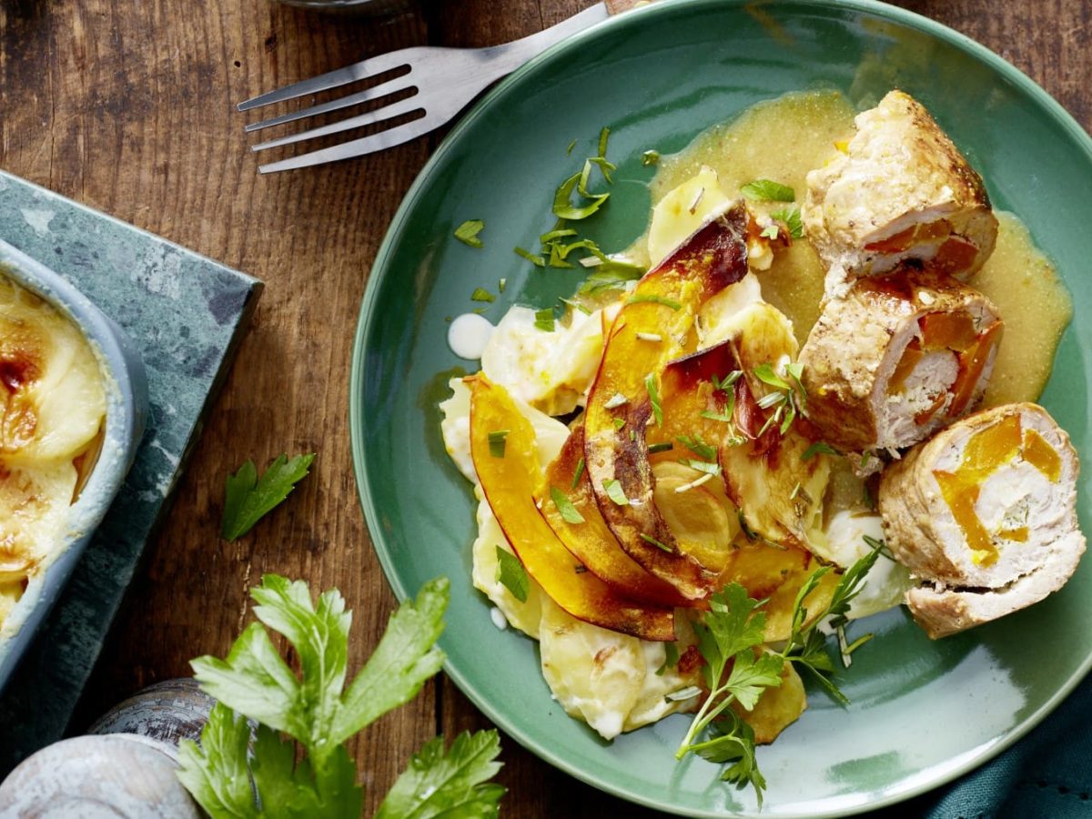 Eine Portion Kürbisröllchen mit Gratin auf einem türkisen Teller auf einem hölzernen Hintergrund. Links davon eine Auflaufform. Oben eine Gabel und unten eine Pfeffermühle und Petersilie.