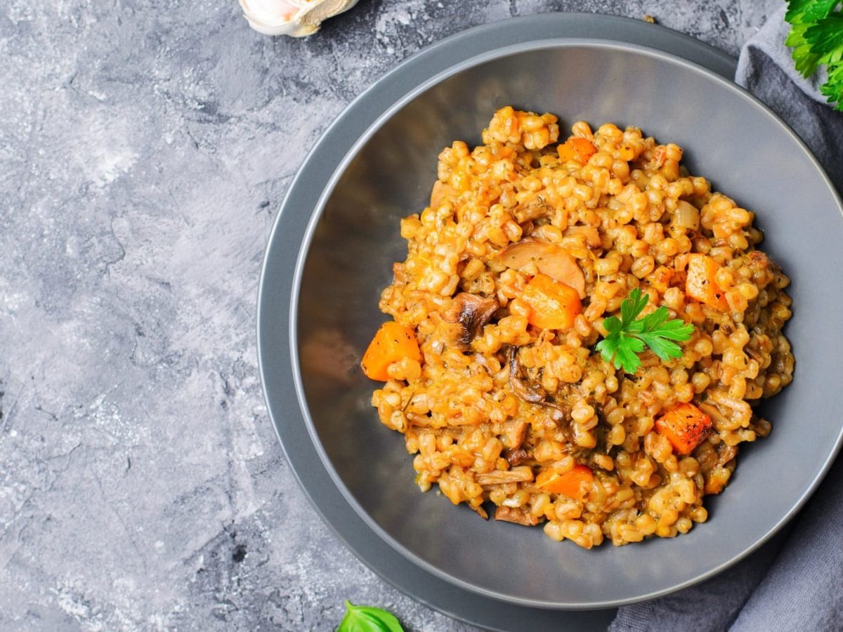 Ein grauer Teller mit dem Risotto mit Kürbis und Pilzen auf grauem Untergrund, mit grauem Tuch und Petersilie.