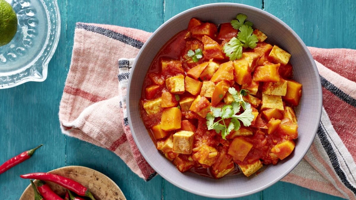 Ein Kürbis-Tofu-Ragout auf einem roten Geschirrtuch auf einem blauen Hintergrund. Links unten Chilischoten und links oben eine Limette.