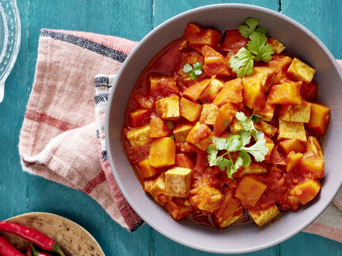 Ein Kürbis-Tofu-Ragout auf einem roten Geschirrtuch auf einem blauen Hintergrund. Links unten Chilischoten und links oben eine Limette.