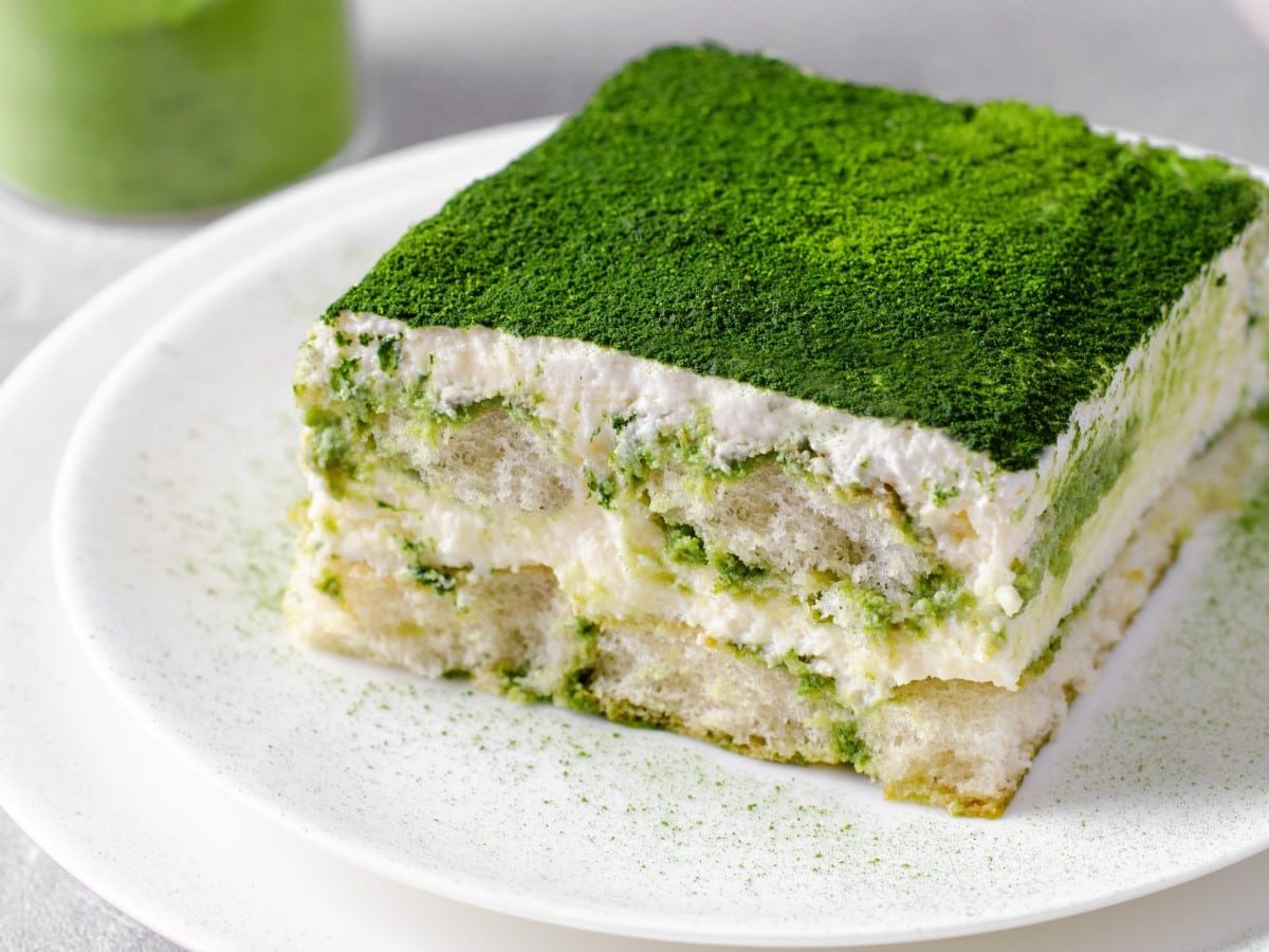 Ein Stück veganes Matcha-Tiramisu auf einem weißen Teller vor einem grauen Hintergrund. Links oben Matcha-Pulver.