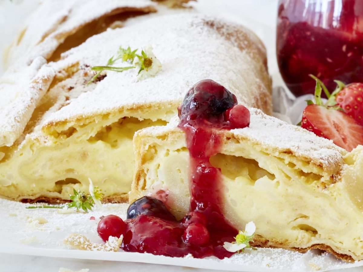 Ein Milchreis-Strudel mit roter Grütze auf einem weißen Servierteller. Rechts davon ein Glas mit roter Grütze.