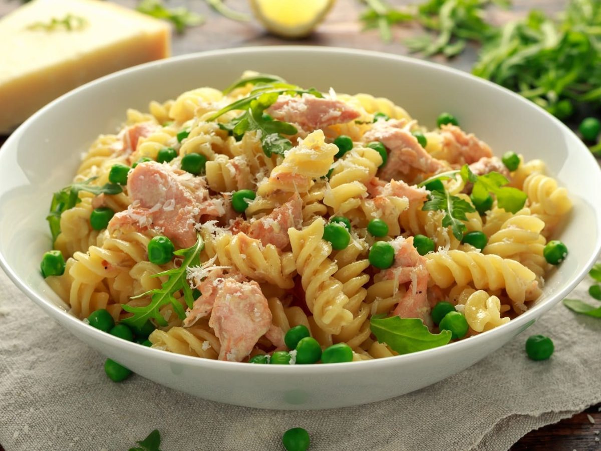 Ein weißer, tiefer Teller mit dem Nudelsalat mit Thunfisch und Erbsen auf heller Serviette, aber dunklem Holztisch. Im Hintergrund sind einige Salatzutaten unscharf zu erkennen. Alles seitlich fotografiert.