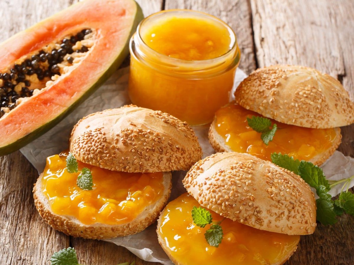 Draufsicht schräg: Ein Glas Papaya-Aufstrich, drei Sesambrötchen mit Papaya-Aufstrich und eine halbe, aufgeschnittene Papaya.