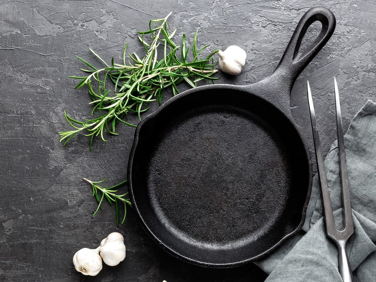 Eine schwarze Pfanne auf dunklem Untergrund mit Rosmarin, Knoblauch und einer Fleischgabel sowie grauer Serviette. Alles direkt von oben fotografiert.