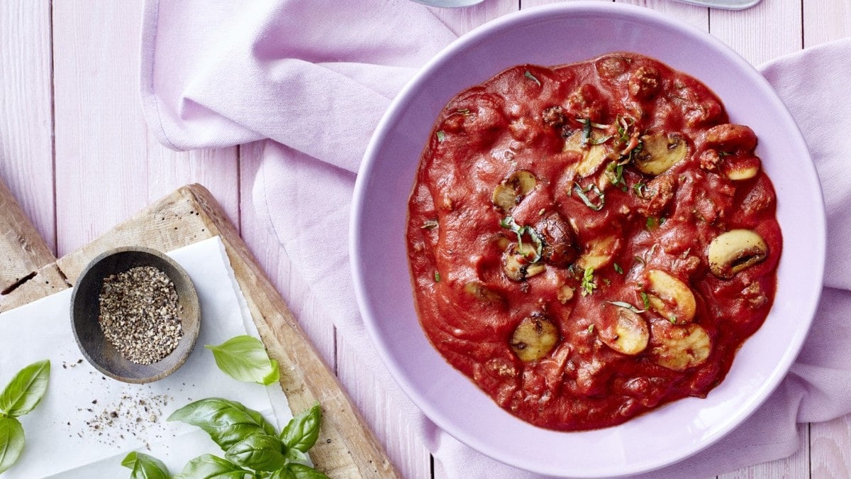 Eine Bolognese mit Pilzen in einem violetten Teller auf einem violetten Geschirrtuch auf einem violetten Hintergrund. Links ein Schneidebrett mit Pfeffer und Basilikum.