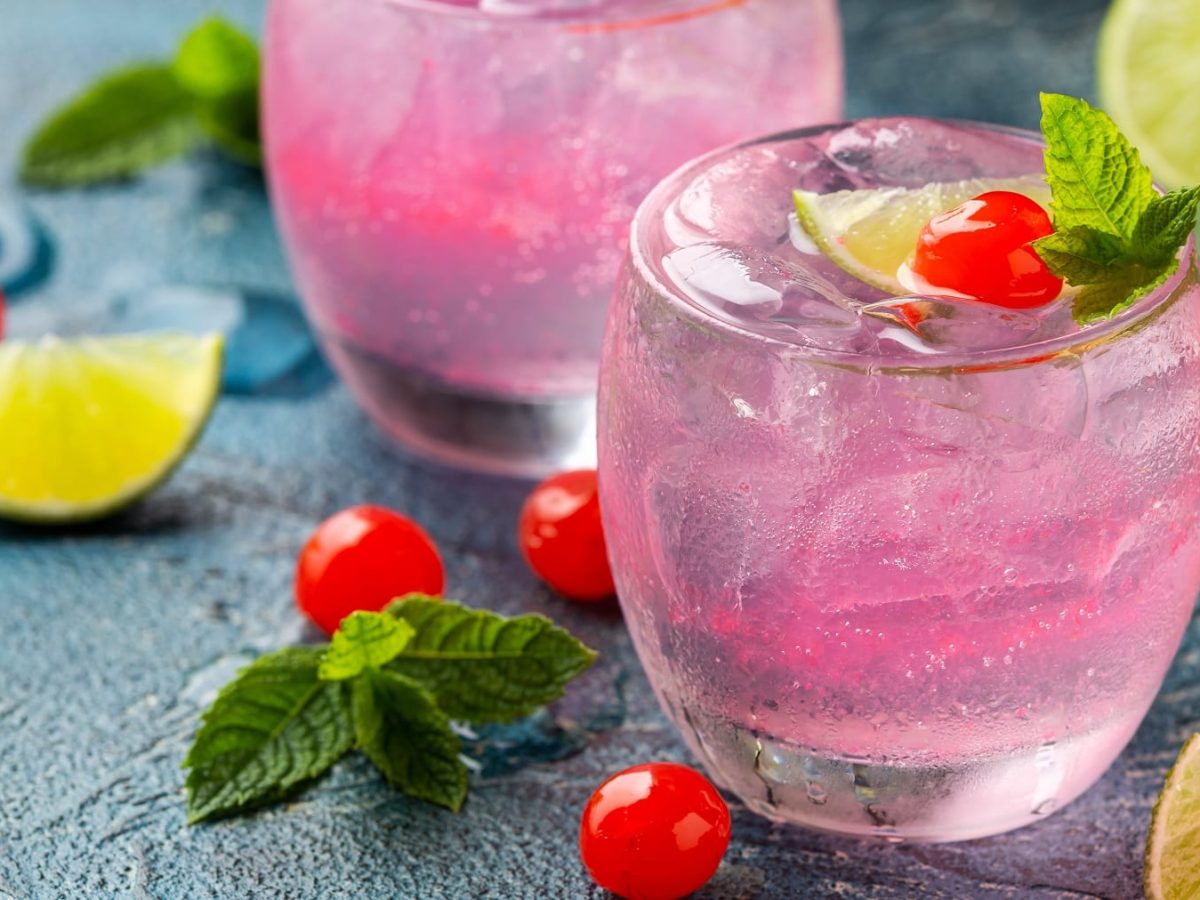 Zwei Gläser mit dem Pink Cherry Gin auf dunkelblauem Tisch mit Limettenspalten und Maraschino-Kirschen dekoriert. Alles seitlich fotografiert.