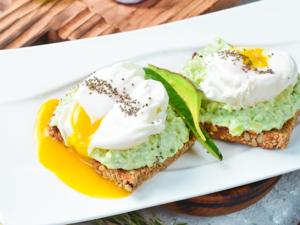 Ein weißer, rechteckiger Teller mit dem pochierten Ei auf Avocado-Feta-Creme und dunklem Brot.