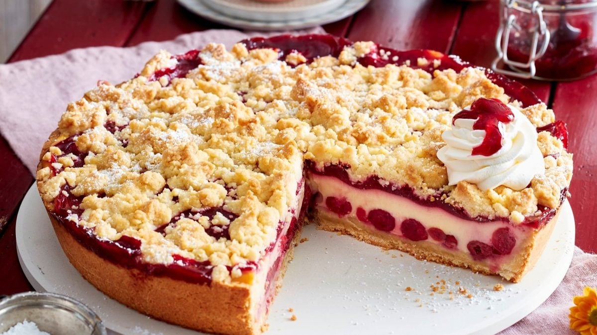 Angeschnittener Pudding-Kirschkuchen mit Streuseln und Sahne auf einem weißen Teller, roter Untergrund und Geschirr und Kirschen im Hintergrund.