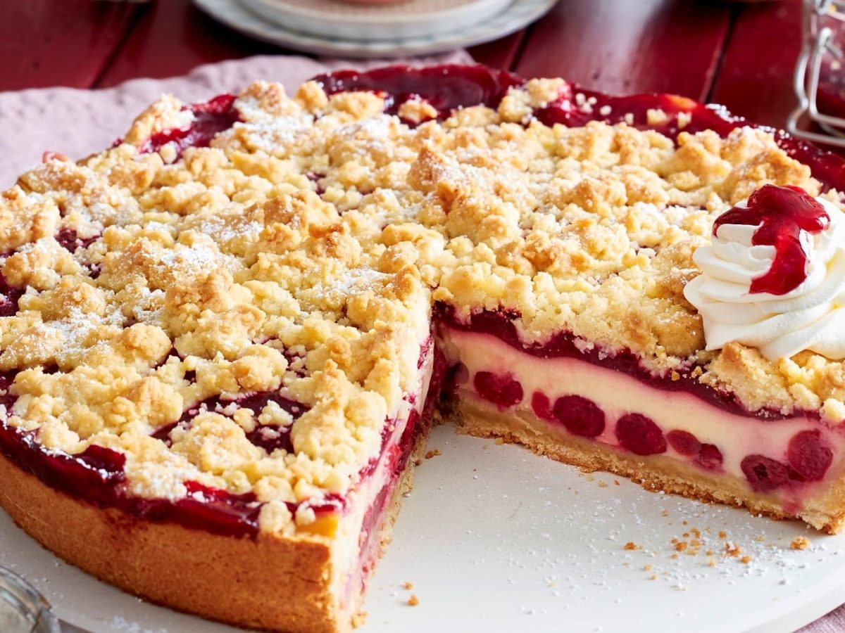 Angeschnittener Pudding-Kirschkuchen mit Streuseln und Sahne auf einem weißen Teller, roter Untergrund und Geschirr und Kirschen im Hintergrund.