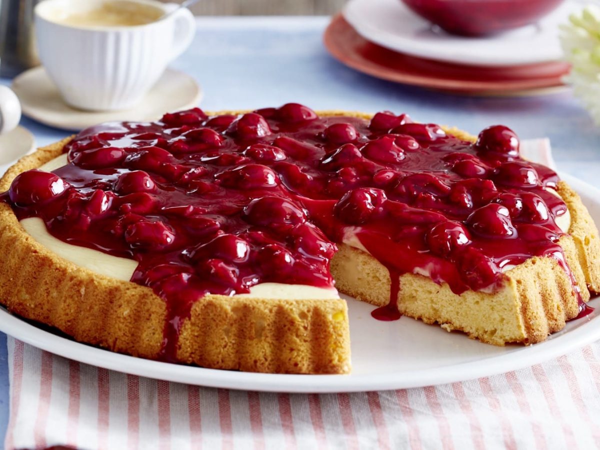 Ein Pudding-Kirschkuchen auf einem weißen Servierteller auf einem weiß-rosa-gestreiftem Geschirrtuch auf einem hellblauem Hintergrund. Links oben Kaffeetassen rechts oben Blumen.