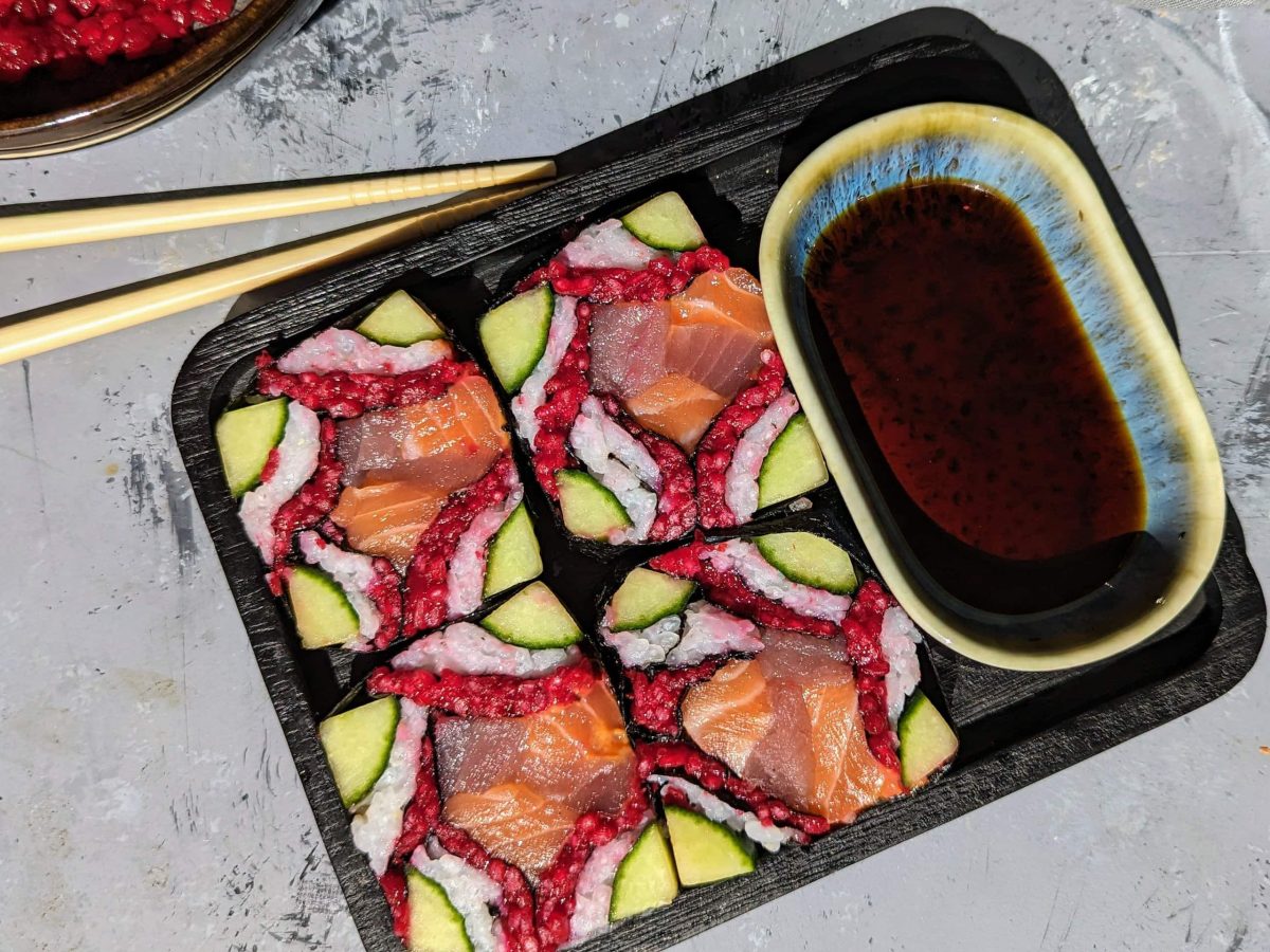 Ein schwarzer viereckiger Teller mit dem Mosaik-Sushi und einer kleinen hellblauen Schale mit Sojasauce von oben fotografiert.