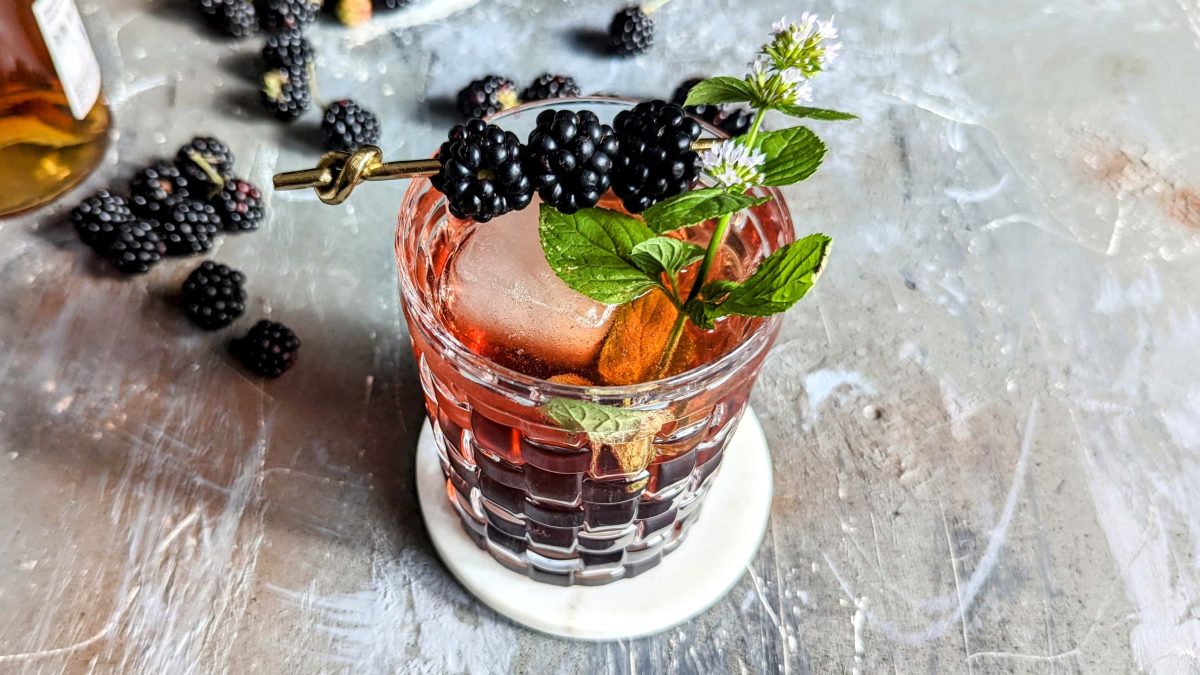 Ein Glas mit dem Brombeer-Smash auf Steinuntergrund. Daneben ein paar Brombeeren und ein kleiner Teil einer Flasche mit Bourbon zu sehen. Alles von oben nach schräg unten fotografiert.