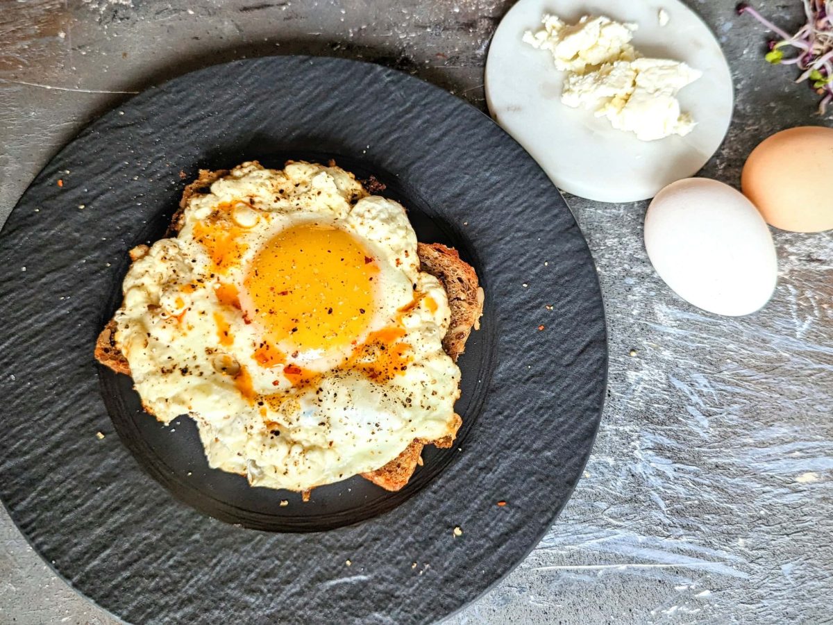 Ein schwarzer Teller mit einer Scheibe Brot und dem Feta-Spiegelei auf Steintisch direkt von oben fotografiert. Rechts oben Feta, Kresse und zwei Eier im Bild.