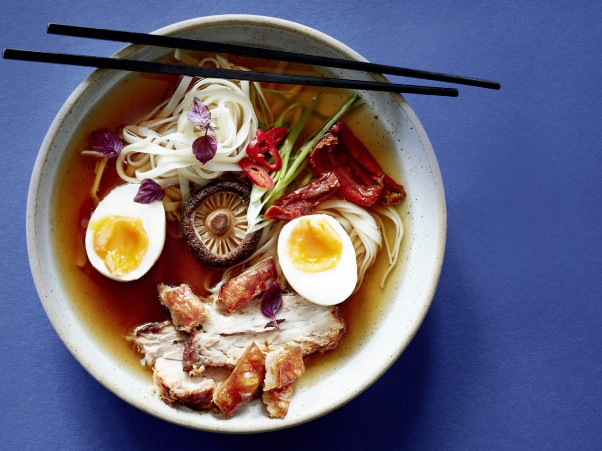 Eine Ramensoup mit Pilz-Dashi und Ei in einer weißen Schüssel vor einem blauen Hintergrund. Auf der Schüssel liegen Stäbchen.