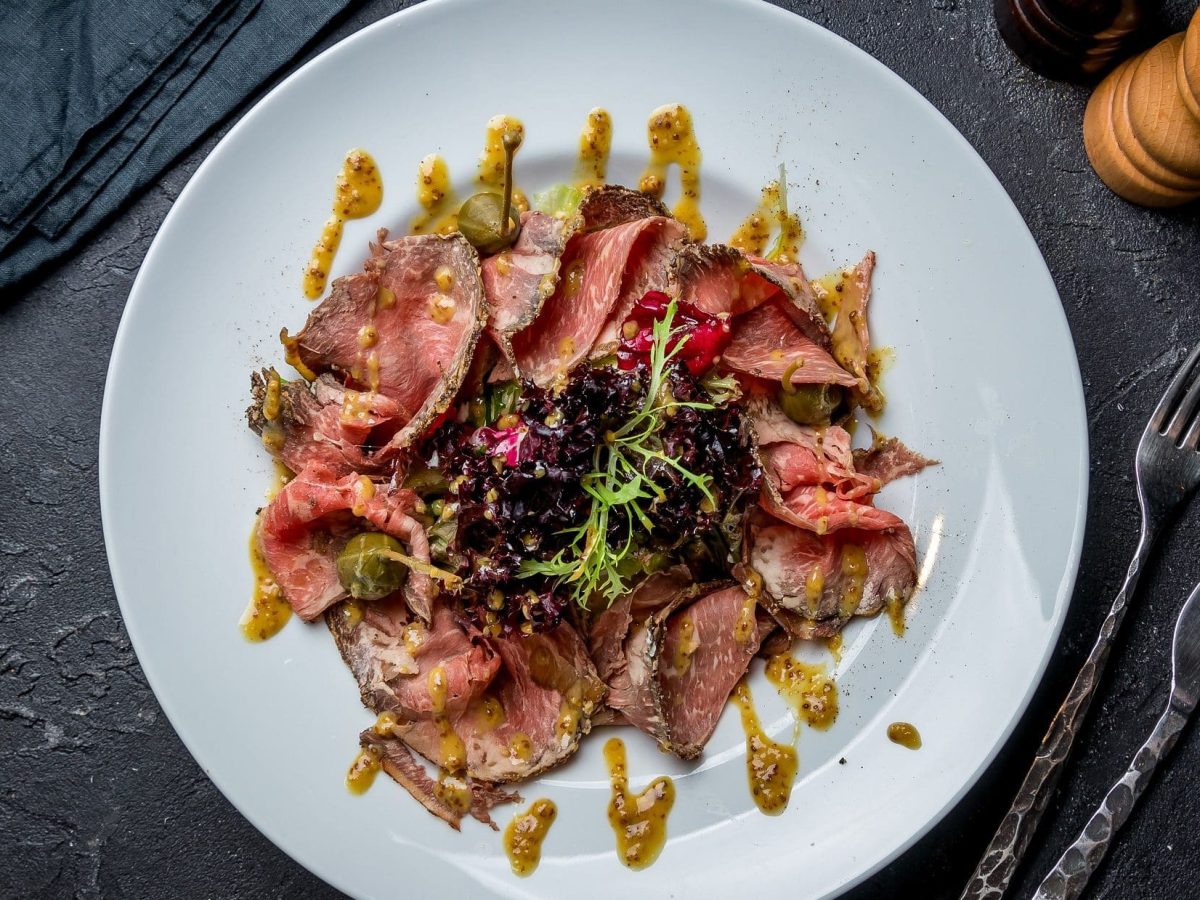 Draufsicht. Auf dunklem Untergrund steht ein weißer Teller mit einem Roastbeef-Salat mit Senf-Dressing. Daneben liegt Besteck.