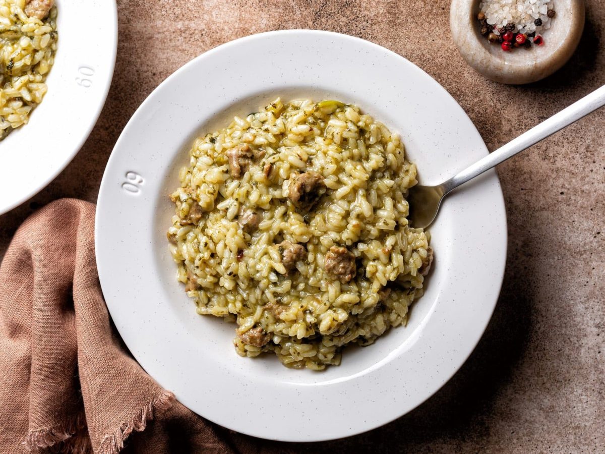 Draufsicht: Untergrund aus Holz mit zwei Tellern Salsiccia-Risotto. Daneben liegt ein Tuch und es steht eine Schale bunter Pfeffer auf dem Tisch.