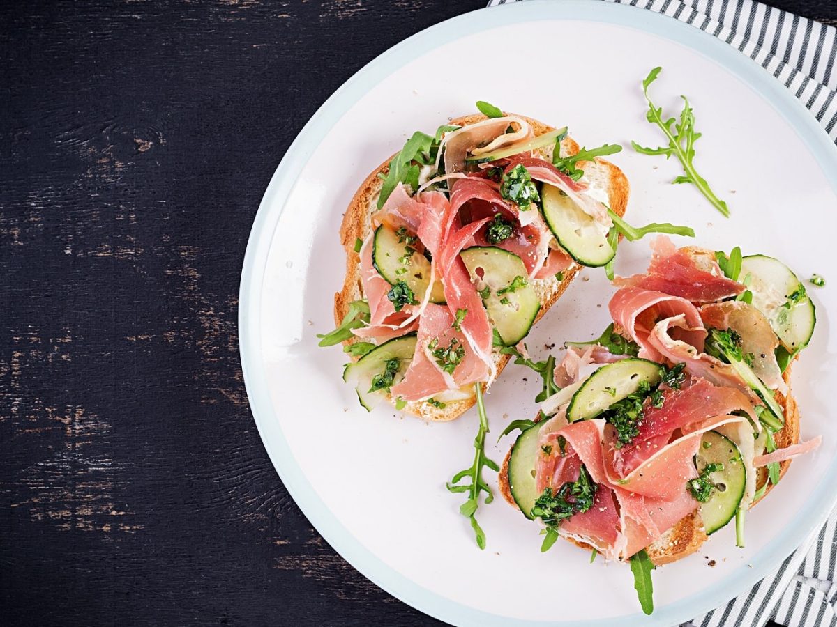 Zwei belegte Brote (Schinke-Gurken-Sandwiches) auf einem weißen Teller von oben fotografiert. Alles auf einem schwarzen Tisch mit weiß-schwarz-Tischdecke.