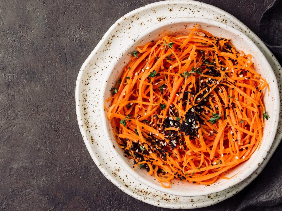 Draufsicht: Dunkler Untergrund und ein weißer Teller mit einem Sesam-Karotten-Salat mit Mandelsauce.