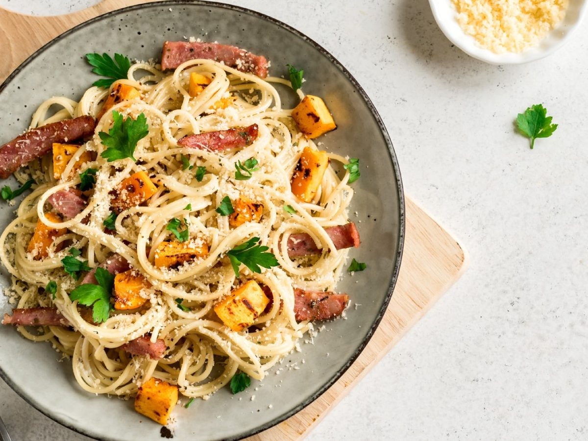 Ein heller Teller mit dunklem Rand und den Spaghetti mit Kürbis und Speck. Alles auf einem weißen Tisch von oben fotografiert.