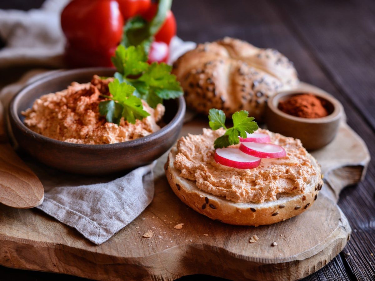 Original Spundekäs nach Winzerart aus Rheinhessen und Hessen in schwarzer Schale und auf Brötchen auf Holz und dunklem Untergrund. Im Bild Radieschen, Paprika und ein Schälchen Paprikapulver. Draufsicht.