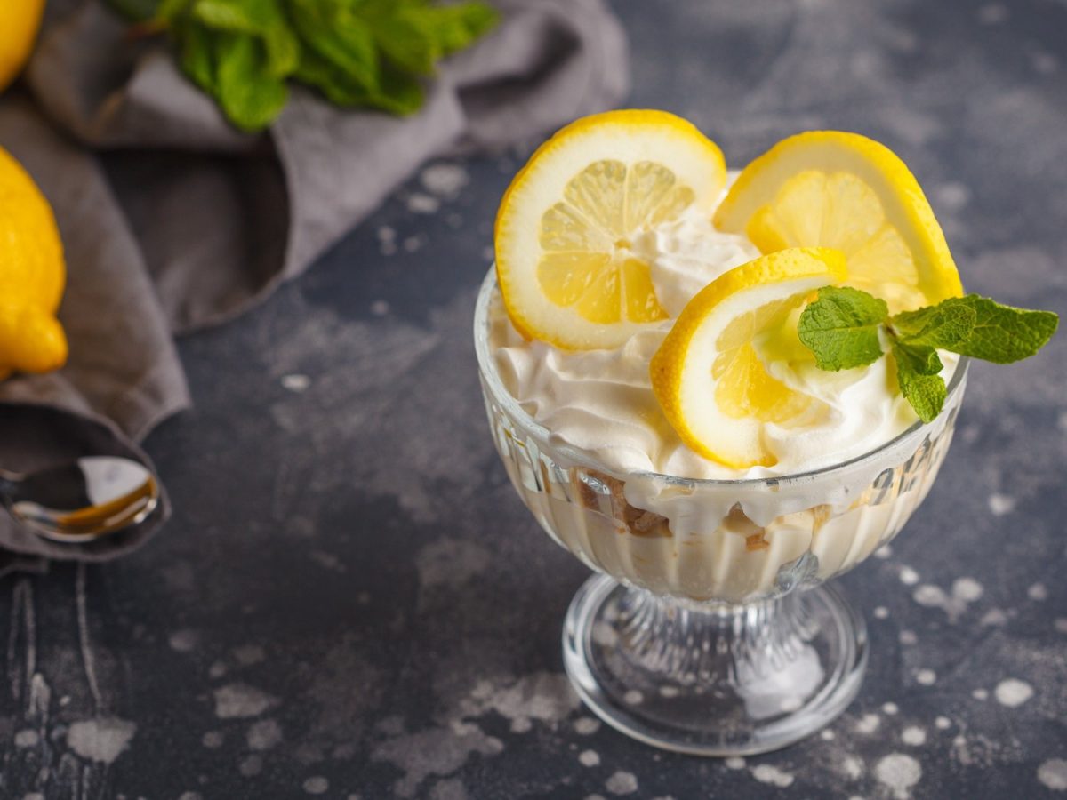 Ein Glas Syllabub, ein weiße Creme aus Zitrone, Weißwein und Sahne, garniert mit Minze und Zitronenscheiben, daneben liegen Zitronen.