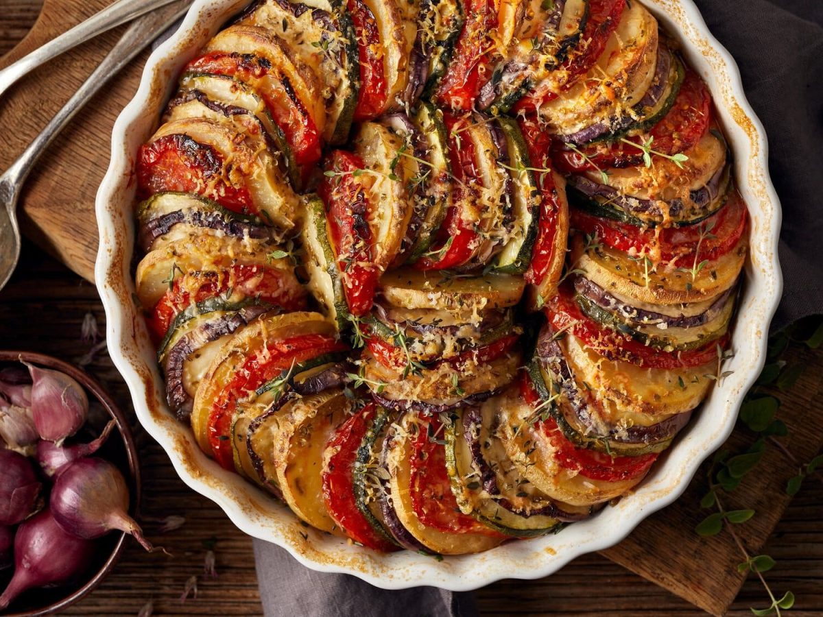 Eine runde Auflaufform Tian, französischer Gemüseauflauf, bei dem Tomaten, Zucchini und Aubergine fächerartig angeordnet werden. Hier in der Draufsicht und mit Käse überbacken, daneben eine Schale mit Schalotten.