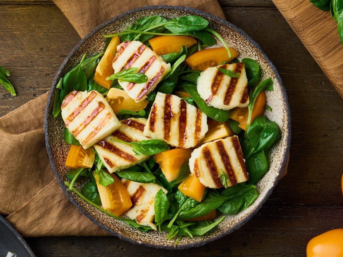 Draufsicht: eine gegrillter Tomaten-Halloumi-Salat auf Holzuntergrund. Daneben liegen gelbe Tomaten und Spinatblätter.