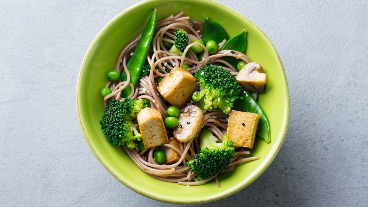 Draufsicht: Eine grüne Schüssel mit vegane Gemüsepasta mit gebratenem Tofu auf hellem Untergrund.