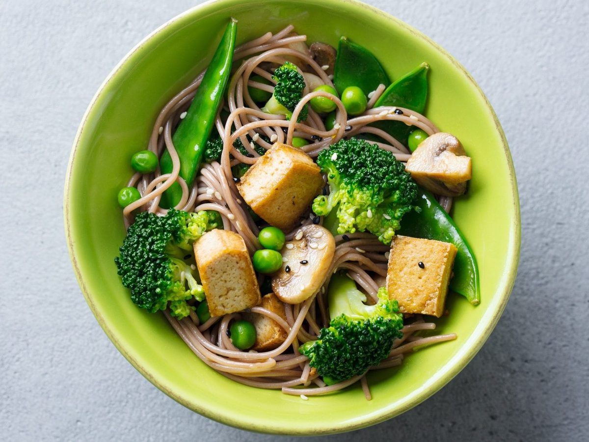 Draufsicht: Eine grüne Schüssel mit vegane Gemüsepasta mit gebratenem Tofu auf hellem Untergrund.