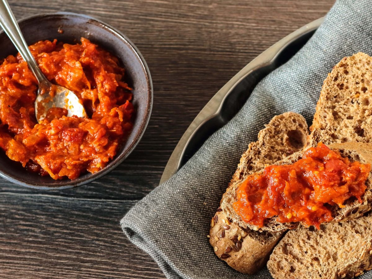 Draufsicht: Dunkler Untergrund, eine dunkle, kleine Schale mit einem veganen Tofu-Tomaten-Aufstrich. Auf einem grauen Tuch liegen mehrere Scheiben Brot.